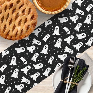 May include: A Halloween-themed table setting with a black and white ghost-patterned table runner. The runner features white ghost illustrations and stars on a black background. A white plate, silverware, and a black napkin are set on the table, along with two pies.