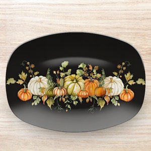 May include: A black oval serving platter with a watercolor design of pumpkins and fall foliage. The pumpkins are in shades of orange, green, and white, with green leaves and sprigs of berries. The platter is a decorative piece for autumn.