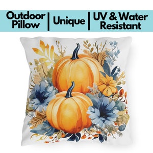 May include: White outdoor pillow with a watercolor design featuring two large orange pumpkins, blue flowers, and autumn foliage. The pillow has the words "Outdoor Pillow | Unique | UV & Water Resistant" printed on it.