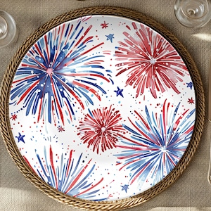 May include: A white ceramic dinner plate with a red, white, and blue watercolor firework design. The plate has a gold rim.