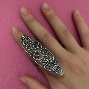 May include: A silver-toned ring with an elongated oval shape. The ring features an intricate, swirling design across its surface. The ring is worn on a finger against a pink background.