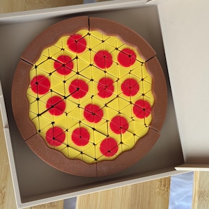 May include: A pizza-shaped puzzle with yellow, red, and brown colors. The puzzle is in a white box and has a brown crust, yellow cheese, and red pepperoni. The pizza is cut into triangular pieces.