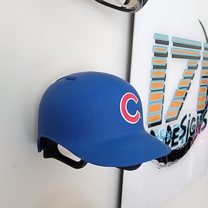 May include: A collection of sports helmets, including a blue baseball helmet with a red and white "C" logo, an orange football helmet, and a gold football helmet. The helmets are mounted on a white wall.