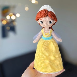 May include: A crocheted doll with a yellow dress, white head covering, and brown hair. The doll is wearing a blue and white beaded necklace.