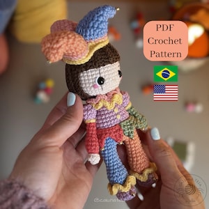 Pode incluir: Uma boneca de crochê com um chapéu de bobo da corte em azul, rosa e amarelo, cabelo castanho e uma roupa multicolorida com seções vermelhas, roxas e azuis. A imagem inclui o texto PDF Crochet Pattern, uma bandeira do Brasil e dos EUA.