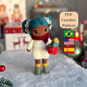 Pode incluir: Boneca de crochê feita à mão com cabelo turquesa, cachecol vermelho e vestido branco. A boneca segura uma cesta vermelha com um pequeno pássaro colorido dentro. A imagem inclui o texto "PDF Crochet Pattern" e bandeiras.