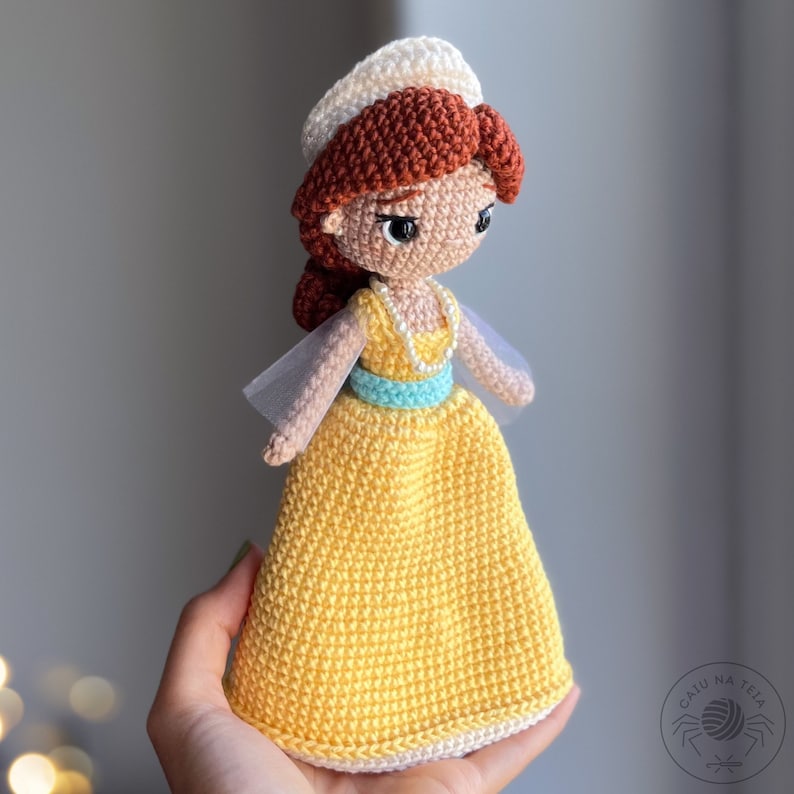 May include: A crocheted doll with a yellow dress, a white crown, and brown hair. The doll has a blue sash and a pearl necklace.
