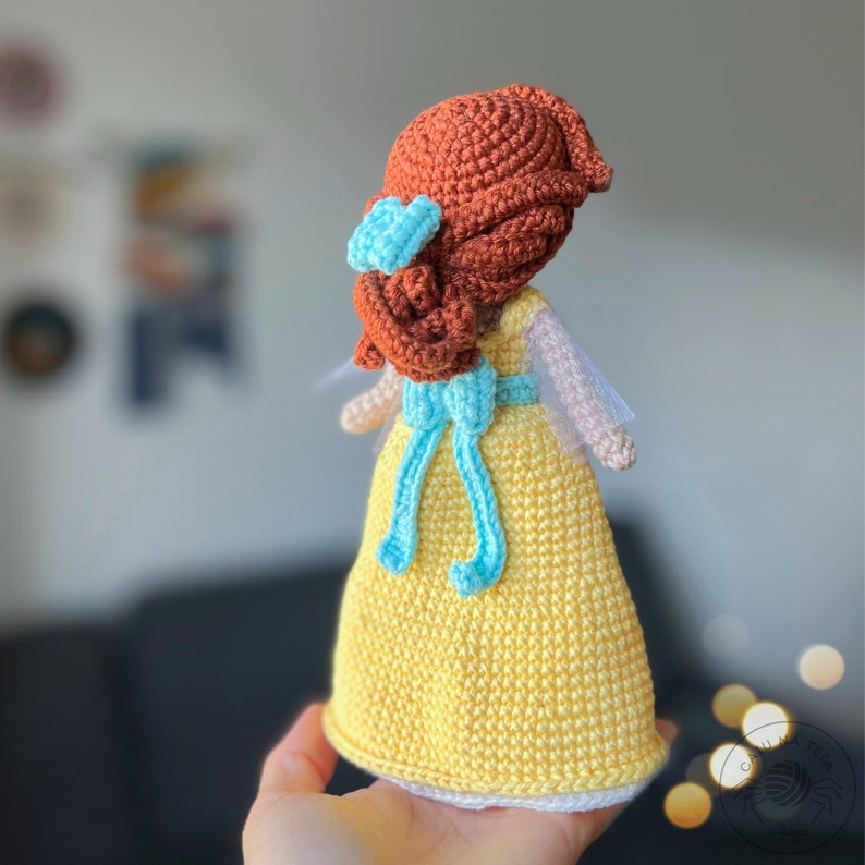 May include: A crocheted doll with brown hair, a yellow dress, and a blue ribbon. The doll is wearing a white crocheted hat.