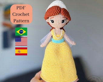 May include: Crochet pattern PDF for a yellow dress with a blue sash and a white hat. The doll has red hair and brown eyes. The pattern is available in Portuguese, English, and Spanish.