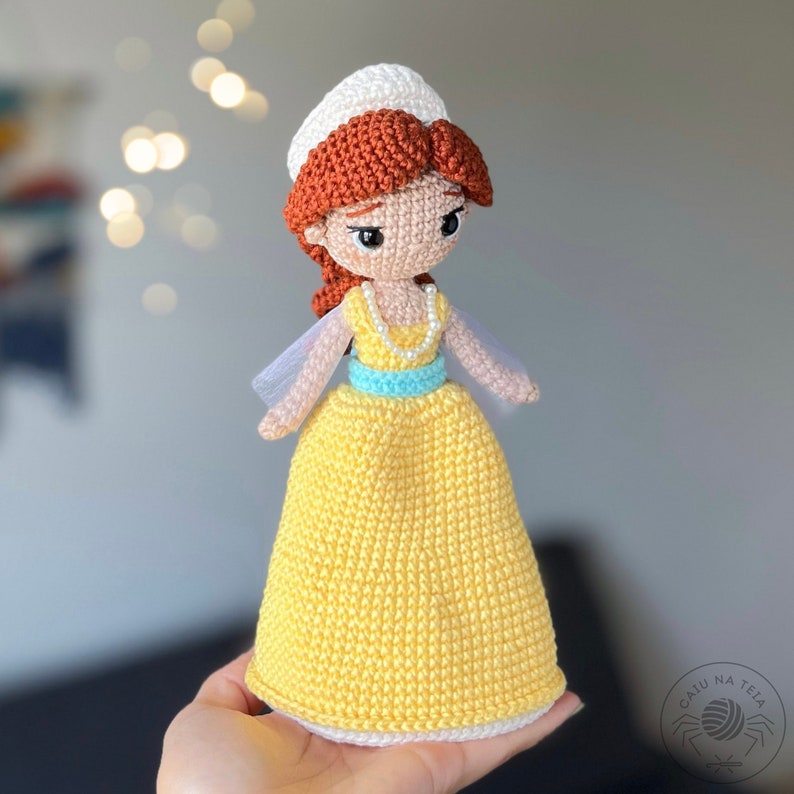 May include: A crocheted doll with a yellow dress, blue sash, and white hat. The doll has red hair and brown eyes. The doll is holding a pearl necklace.