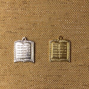 May include: Two pendants, one silver and one gold, each shaped like an open book with Hebrew text. The pendants have a decorative border and a small loop at the top for a chain. The background is a textured, golden-brown fabric.