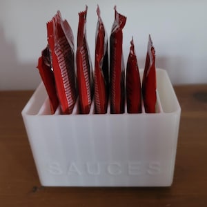 Pode incluir: Um organizador retangular de plástico branco com ranhuras verticais contendo vários pacotes de condimentos vermelhos. A palavra SAUCES está gravada em letras maiúsculas na parte frontal do recipiente, que apresenta uma textura canelada.