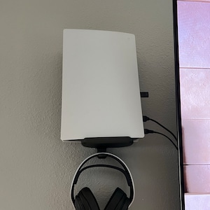 May include: A white gaming console mounted on a wall with a black headset hanging from the console.