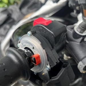 May include: Close-up of a motorcycle handlebar control assembly. The assembly features a black grip, a red switch, and a clear plastic ring. The components are mounted on a motorcycle, with other parts visible in the background.