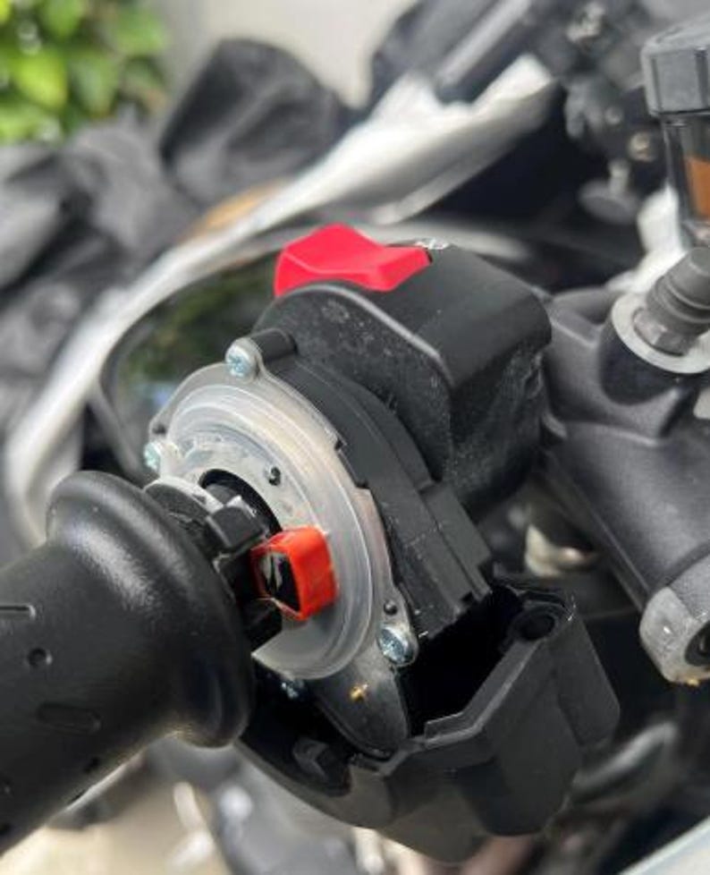 May include: A close-up of a motorcycle handlebar control assembly. The assembly includes a black grip, a red switch, and a clear plastic ring. The components are mounted on a motorcycle, with other parts visible in the background.
