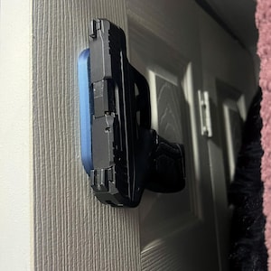 May include: A black handgun with a black grip is mounted on a blue holder, attached to a white door. The gun is positioned horizontally, with the barrel facing left. The door has a vertical wood grain texture.