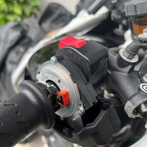 May include: Close-up of a motorcycle handlebar control assembly. The assembly features a black grip, a red button, and various mechanical components. The image highlights the details of the motorcycle's controls.