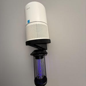 Puede incluir: Un purificador de aire blanco con una base negra está montado en una pared, combinado con un matamoscas negro. El purificador de aire tiene un diseño cilíndrico con una sección inferior perforada. El matamoscas tiene una jaula cilíndrica con luz azul.