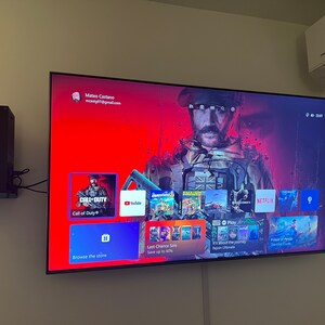 May include: A television screen displaying a video game console menu with a red background. The menu includes icons for various apps and games, including Call of Duty, YouTube, Netflix, and Play. The text "Browse the store" and "Last Chance Sale Save up to 50%" is visible on the screen.