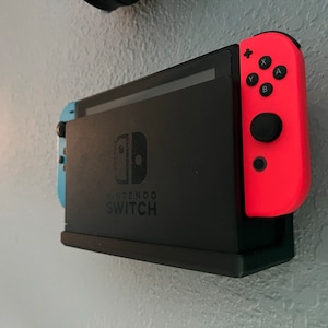 May include: A black Nintendo Switch console with a red Joy-Con controller attached. The console is mounted on a wall with a white background.