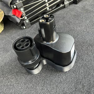 Puede incluir: Adaptador de carga para vehículos eléctricos negro con un conector redondo y una base rectangular más grande. El adaptador tiene tres clavijas y un puerto circular. El fondo incluye un carro plegable negro.