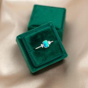 May include: A silver ring with a rectangular, light blue opal center stone. The ring features small, leaf-like accents on either side of the opal. The ring is displayed in a dark green velvet ring box.