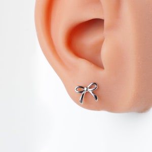 May include: A silver bow earring, shown in a close-up view of a person's earlobe.