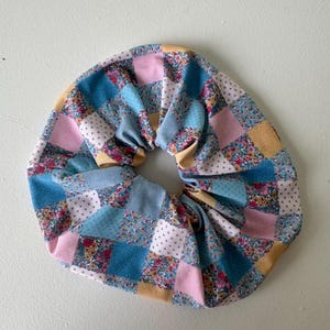 May include: A large, handmade scrunchie with a patchwork design. The fabric features squares of blue, pink, yellow, and white, each with floral or dotted patterns. The scrunchie is designed to hold hair and is made of soft fabric.