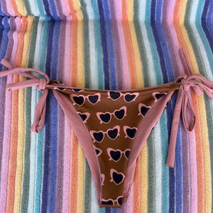 May include: Brown bikini bottom with a heart-shaped sunglasses pattern in black and pink. The bikini bottom has pink ties on each side. The background is a colorful striped towel.