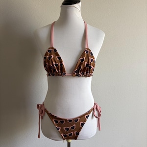 May include: A two-piece bikini set displayed on a mannequin. The top is a triangle style with pink straps and a brown fabric patterned with dark purple hearts. The bottoms are brown with the same heart pattern and pink tie sides.