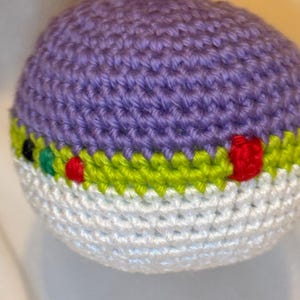 May include: A handmade crochet ornament in a spherical shape. The top is purple, the middle section is white with a green and yellow stripe, and the ornament has red, green, and black accents. A purple loop is attached for hanging.