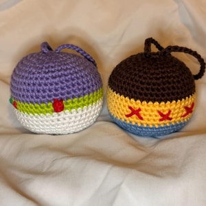 May include: Two handmade crochet ornaments. One is purple, green, white, and red. The other is brown, yellow, and blue, with red cross-stitch details. Each has a loop for hanging.