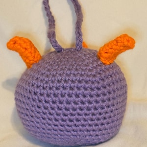 May include: A handmade crochet ornament in the shape of a round creature. The body is a solid lavender color, with two orange horns and a lavender loop for hanging. The ornament is made with a tight crochet stitch.