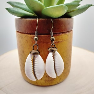 May include: A pair of white shell earrings with gold wire accents hanging from bronze hooks. The earrings are displayed against a wooden background.