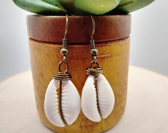 May include: A pair of white shell earrings with gold wire accents hanging from bronze hooks. The earrings are displayed against a wooden background.