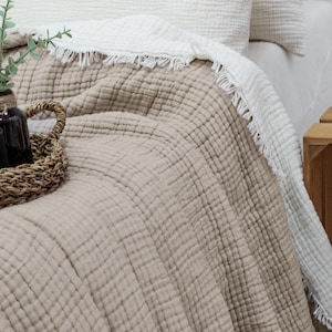 Könnte beinhalten: Ein Bett mit einer beigefarbenen, strukturierten Decke und weißen Kissen. Ein geflochtener Korb enthält eine Tasse, eine kleine Tasse und eine dunkle Flasche. Eine weiße Decke mit Fransen ist über die beige Decke gefaltet. Eine Holzkiste enthält eine Lampe und eine Vase mit Grün.