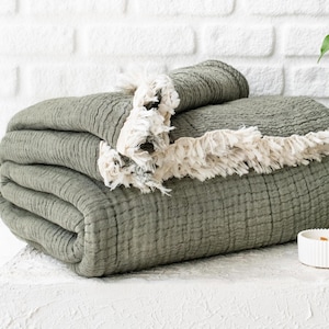 May include: A folded sage green blanket with a textured surface and a cream-colored fringe. The blanket is stacked on a white surface, with a white brick wall in the background. A small white candle holder is also present.