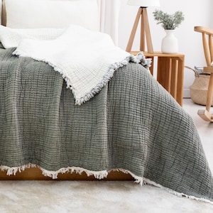 May include: A bed is dressed with a sage green textured blanket with a white fringe. White pillows and a white blanket with a fringe are on the bed. A wooden lamp and a rocking chair are in the background.