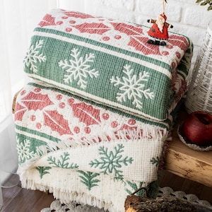 May include: A folded white, green, and pink throw blanket with a snowflake and holly pattern. The blanket has a fringe border and is folded on a wooden surface.