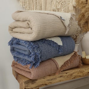 Puede incluir: Tres mantas de algodón apiladas en un banco de madera. La manta superior es beige, la manta del medio es azul y la manta inferior es de color marrón claro. Las tres mantas tienen un borde de flecos.