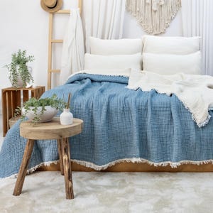 May include: A bed is dressed with a blue textured blanket with a white fringe. White pillows and a white fringed blanket are on the bed. A small wooden table and crate are in front of the bed. A macrame wall hanging is above the bed.