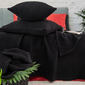 Może przedstawiać: Czarny koc wafelkowy i poduszki na łóżku. Koc jest złożony i drapowany na poduszkach. Poduszki są ułożone jedna na drugiej.