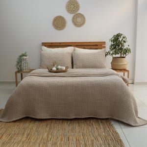 May include: A beige waffle-weave bedspread and matching pillow set on a wooden bed frame. The bed is made up with a woven rug at the foot. Decorative woven wall hangings and potted plants add to the room's decor.