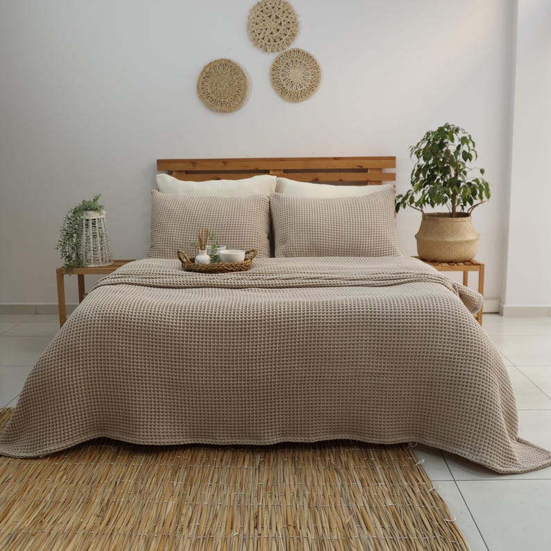 May include: A beige waffle-weave bedspread and matching pillow set on a wooden bed frame. The bed is made up with a woven rug at the foot. Decorative woven wall hangings and potted plants add to the room's decor.