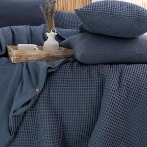 May include: A bed is made with a blue waffle-weave blanket and matching pillows. A wooden tray holds a white ceramic vase, a cup, and dried lavender. The bedding is a solid, muted blue color.