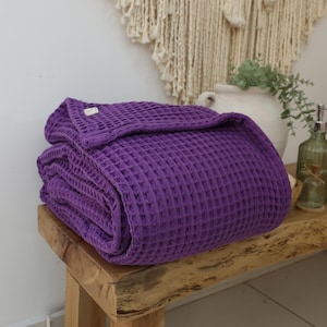 May include: A purple waffle weave throw blanket folded on a wooden bench. The blanket is soft and textured.