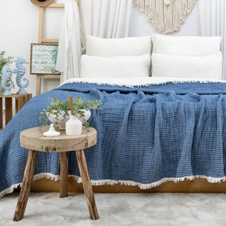 May include: A blue bedspread with a textured weave and a white fringe is draped over a bed with white pillows. A small wooden stool with a natural finish holds white vases and greenery. Decorative seahorse statues are visible.