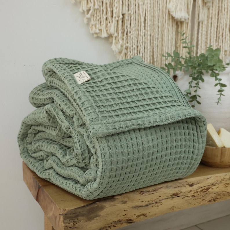 May include: A folded sage green waffle weave throw blanket with a small white tag that says "The Little Market" on a wooden surface.