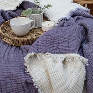 May include: A reversible blanket in lavender and cream with a fringed edge. A woven tray holds a white teacup, a small white candle, and a white wicker basket with greenery. The blanket is draped over a wooden surface.