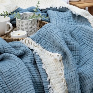 May include: A blue and white cotton blanket with a textured, crinkled appearance. The blanket has a fringed edge and is draped over a tray with a white mug, candle, and small basket with greenery.